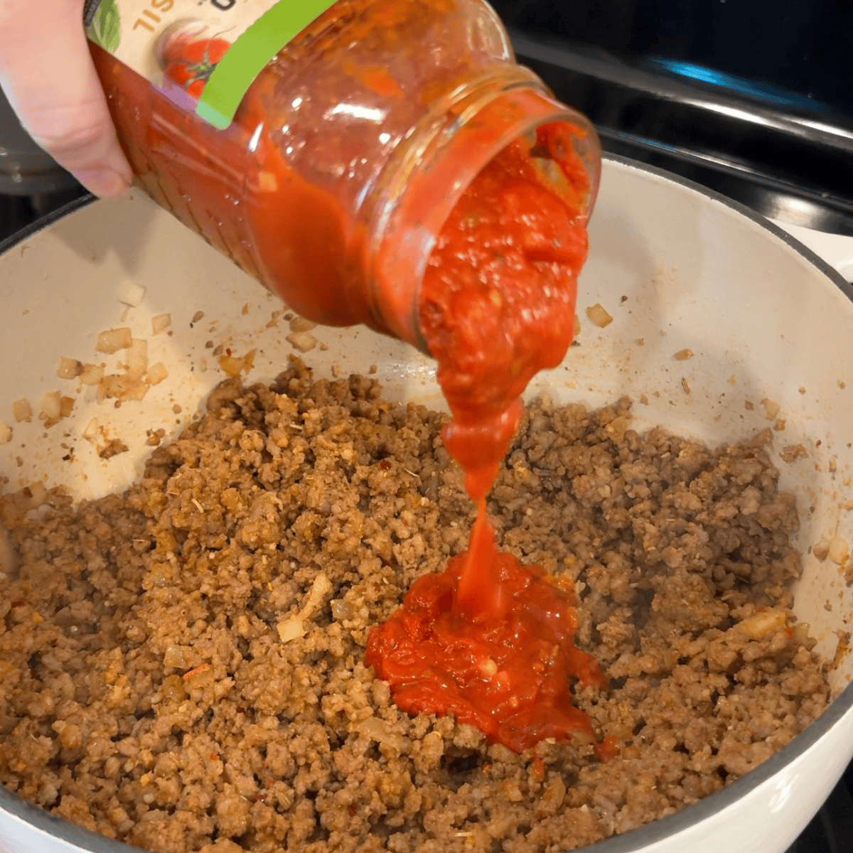 large pot with ground beef and diced onion. hand over pot holding jar of marinara sauce pouring into pot