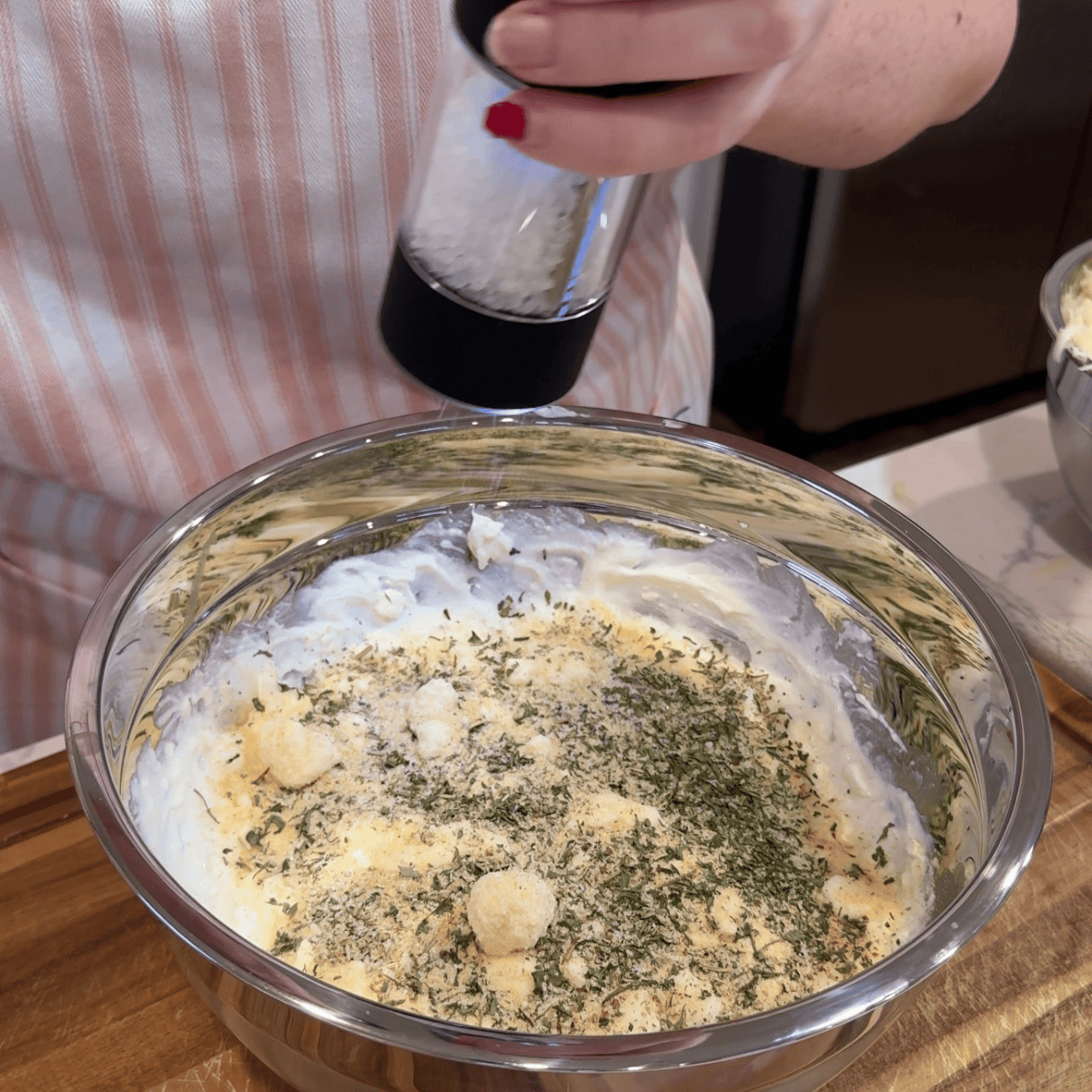 mixing bowl with cheese and seasonings. hand over bowl adding in salt. girl in background with pink  apron