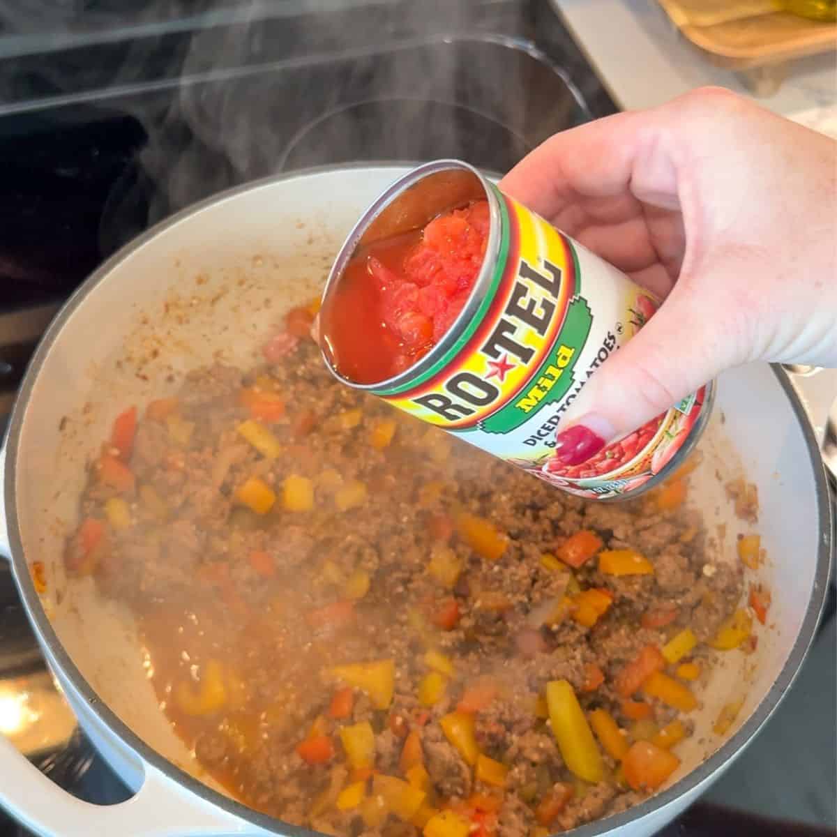 dutch oven with ground beef, and vegetables cooking. hand over dutch oven with can of rotel tomatoes pouring into pot