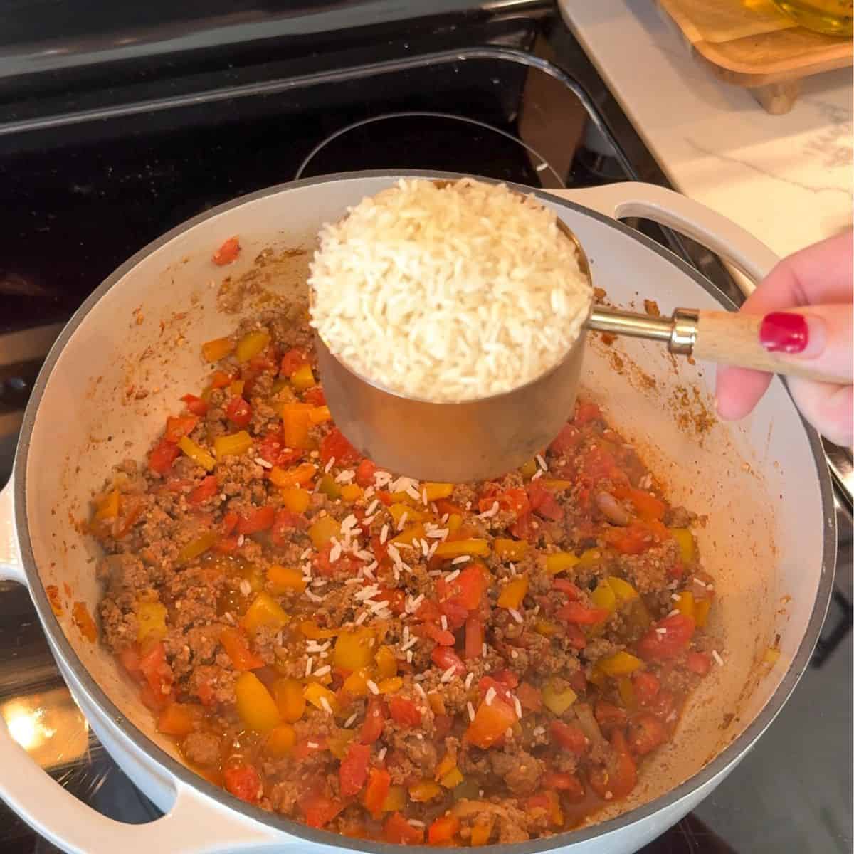 dutch oven with ground beef, bell peppers, and onions cooking. hand over pot with cup of rice pouring into pot