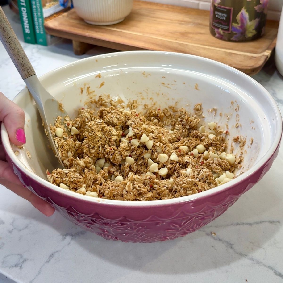 white chocolate no bake energy bites ingredients mixed together in mixing bowl. spatula in bowl with hand on side 