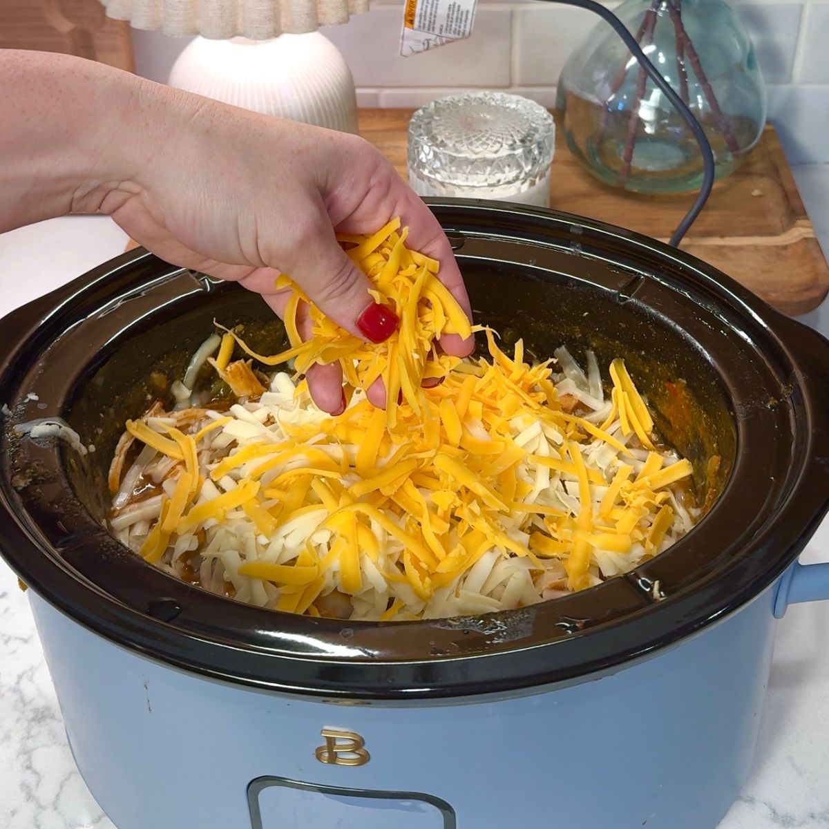hand pouring shredded yellow and white cheese in crockpot