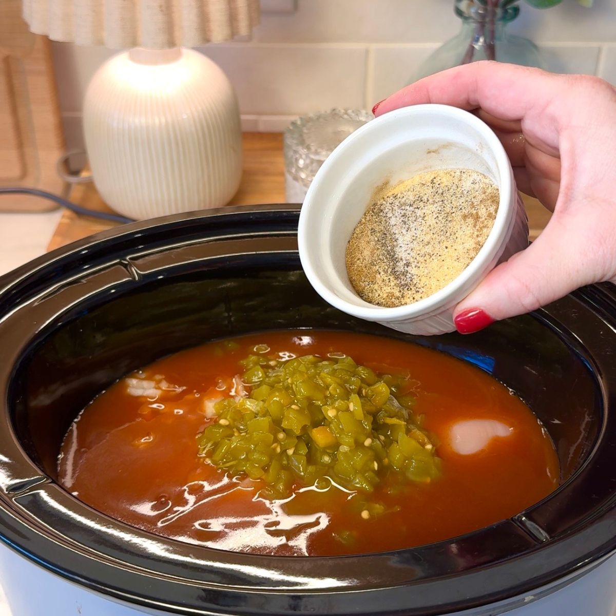 hand holding bowl of seasonings to pour over chicken mixture with sauce and green chilies in crockpot