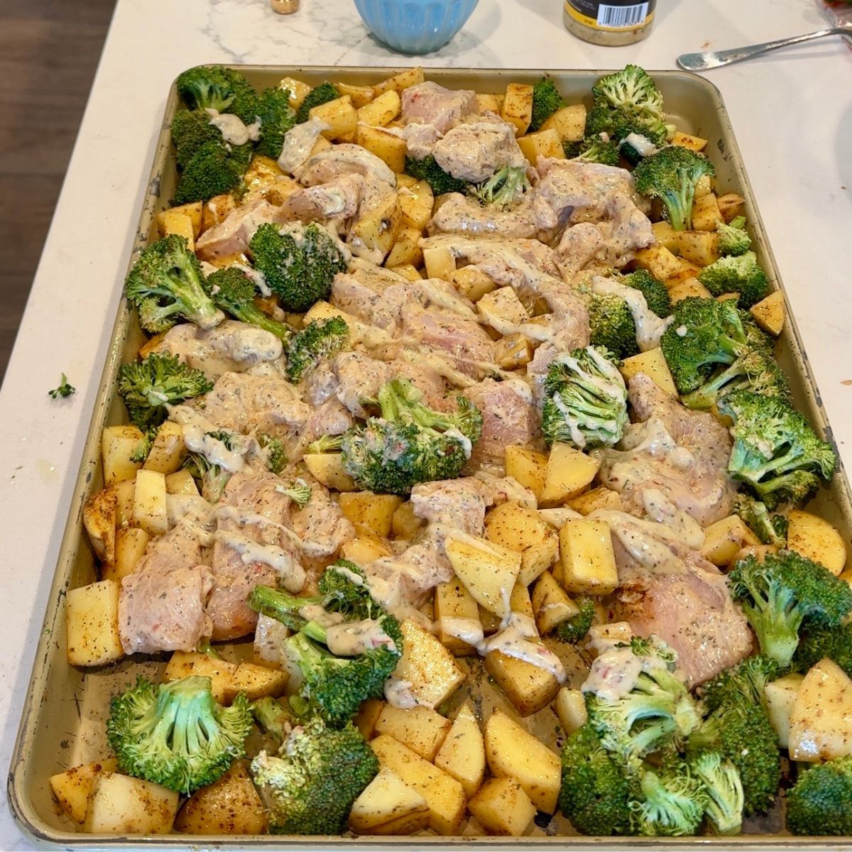 uncooked chicken, potatoes, broccoli on pan with sauce