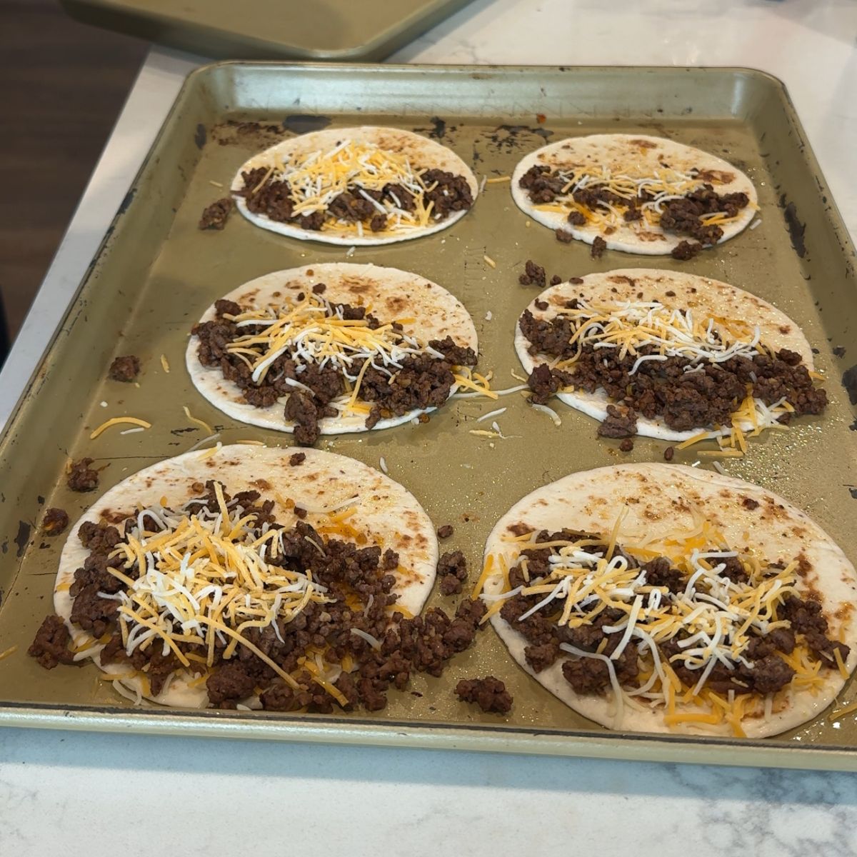 cheese layer added on top of ground beef tacos on baking tray.