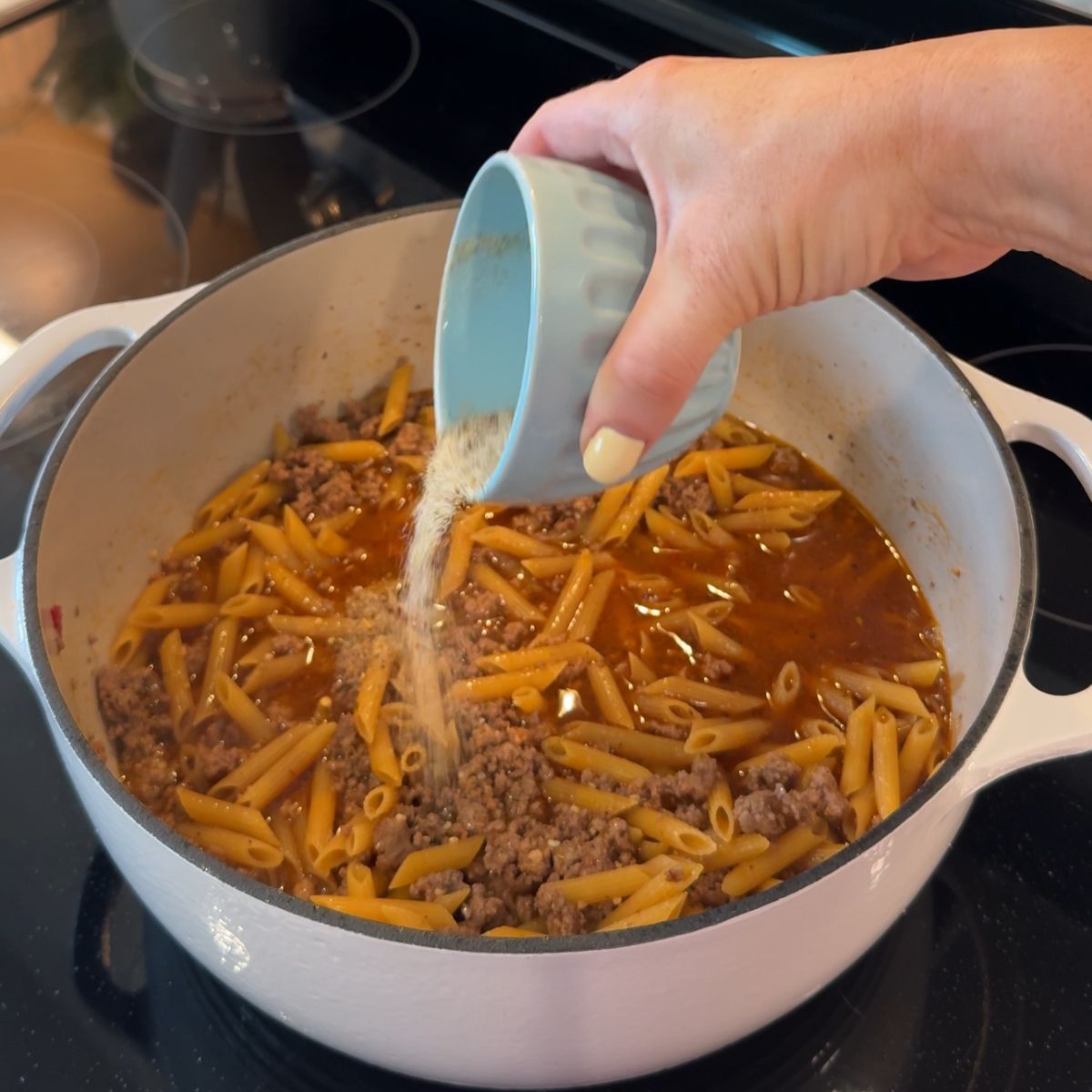 blue cup pouring seasoning into pot with beef and pasta