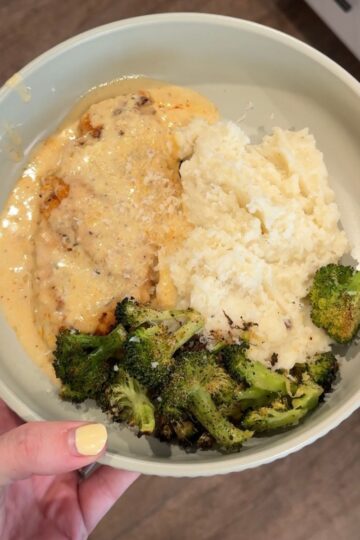 smothered cowboy butter chicken with broccoli, mashed potatoes on green plate with hand on left side