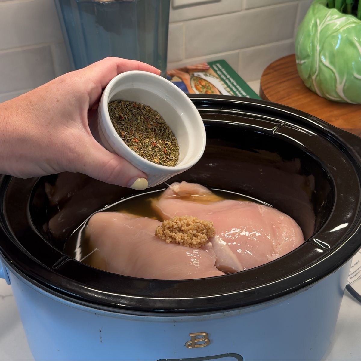 hand holding cup of seasonings over chicken in slow cooker