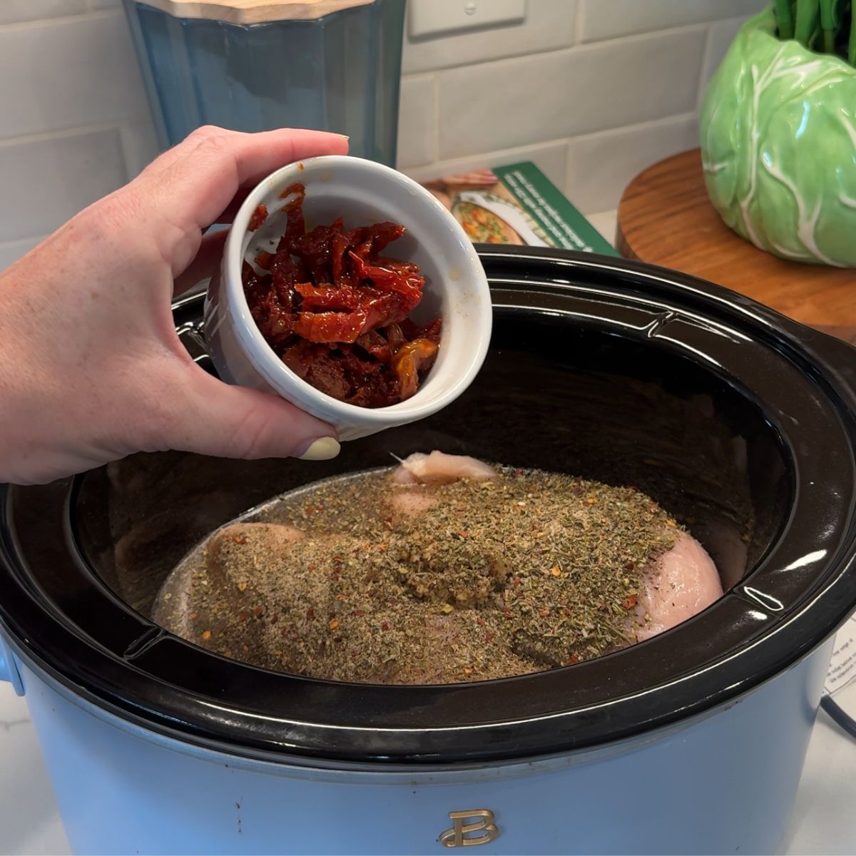 hand holding cup of sun dried tomatoes over slow cooker with seasonings and chicken