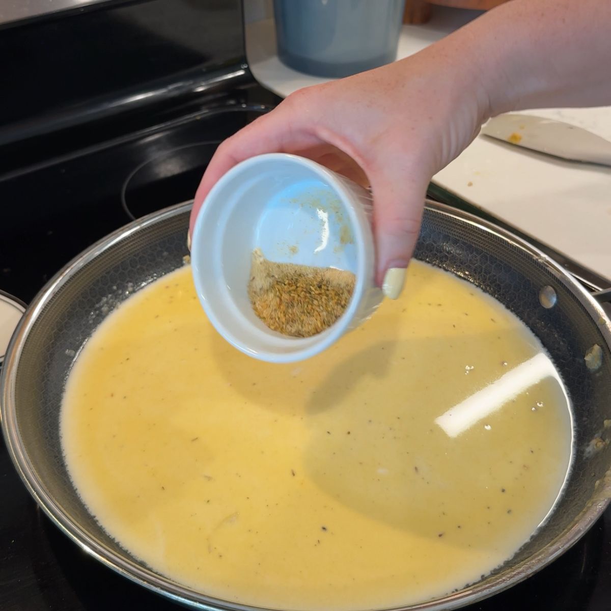 hand holding seasoning in cup over cream sauce in skillet 