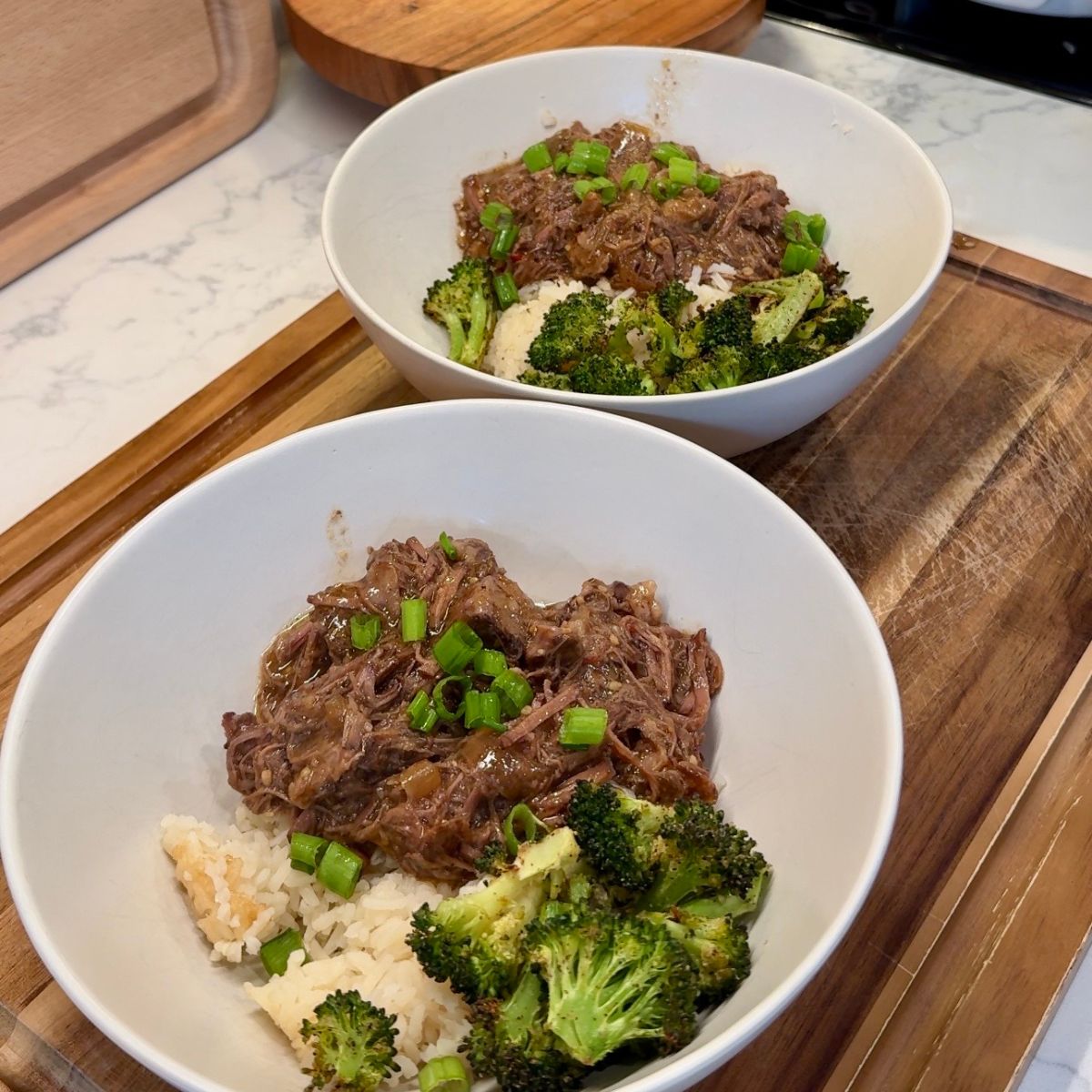 two bowls of slow cooker korean beef bowls with broccoli and rice on wooden board and lamp in background