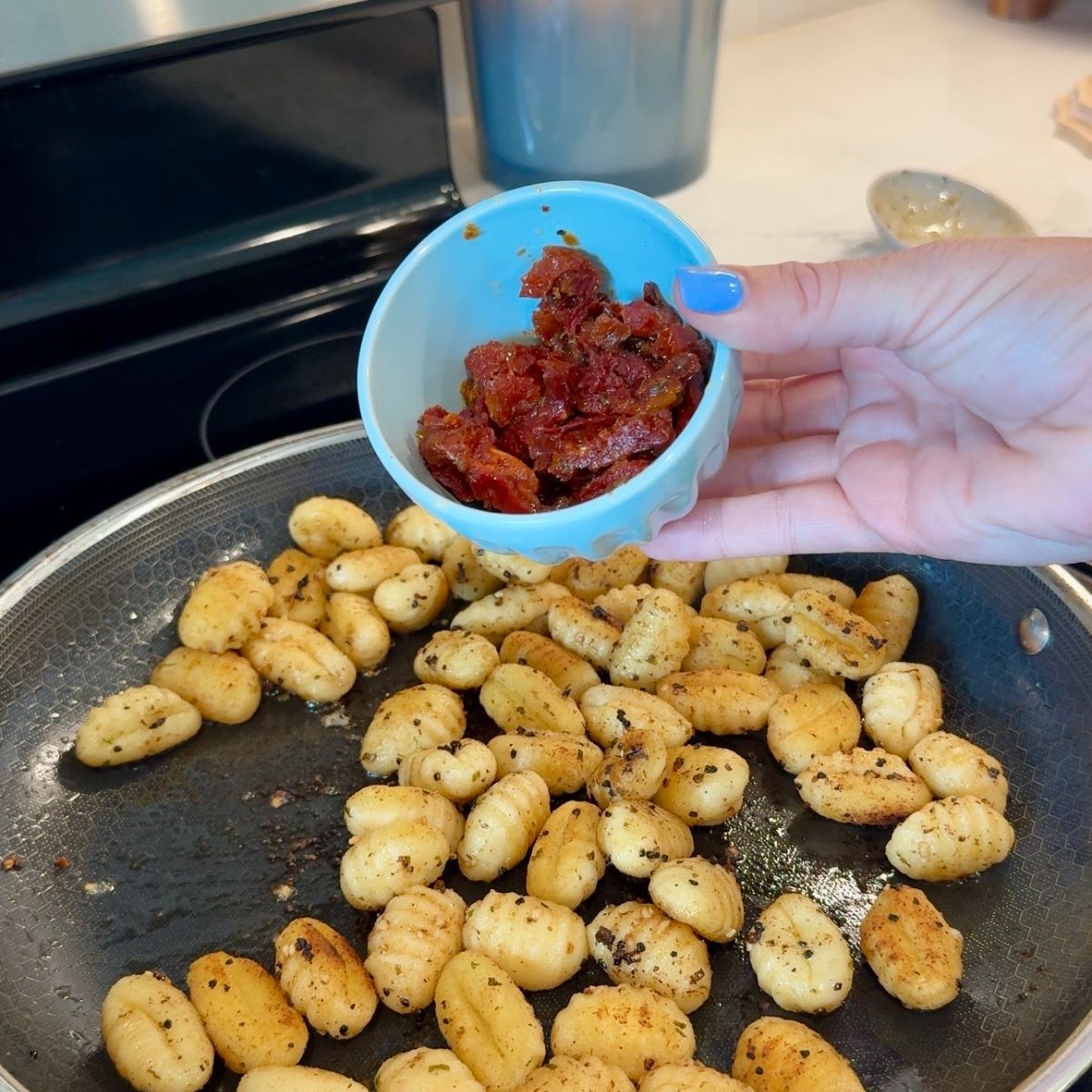 hand holding bowl with sun dried tomatoes over gnocchi in skillet