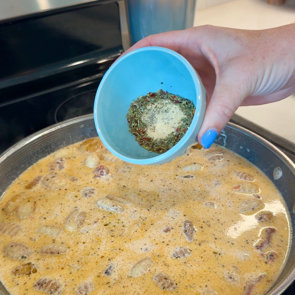 hand holding seasonings in blue bowl over skillet with sauce