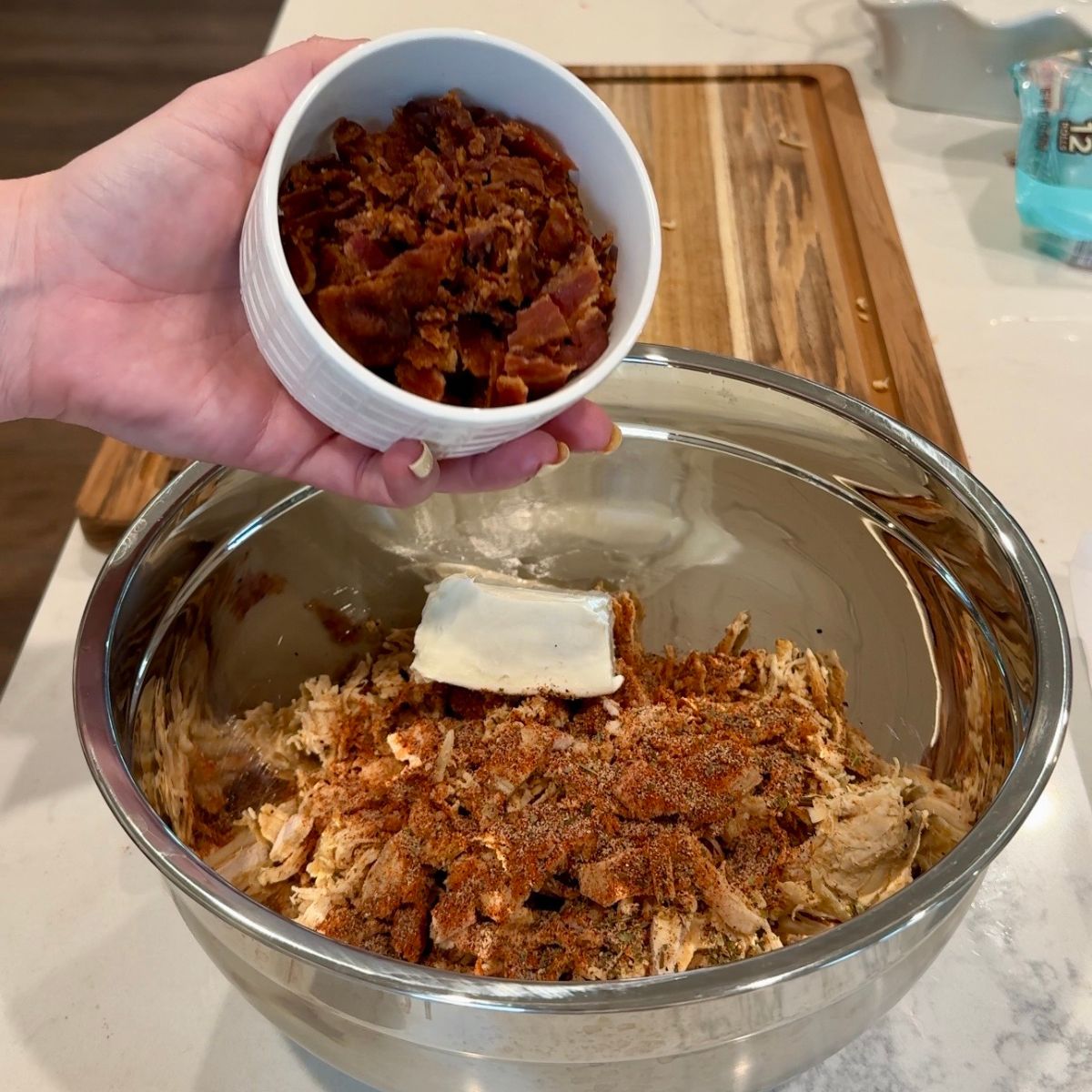 chicken in bowl with cream cheese, and bacon bits in cup that hand is holding