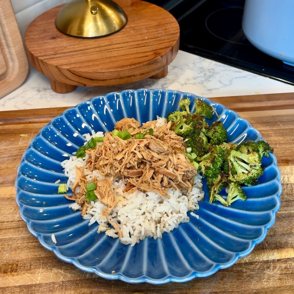 chicken, rice and broccoli on blue plate with green onions on top. wooden board underneath plate