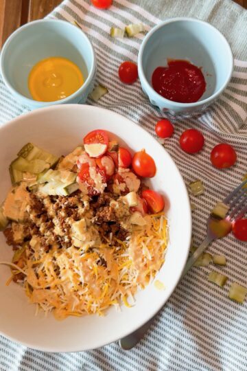 A white bowl with shredded cheese, ground beef, diced pickles, chopped lettuce, cherry tomatoes, and sauce sits on a striped cloth near bowls of ketchup and mustard, with scattered cherry tomatoes and pickles.