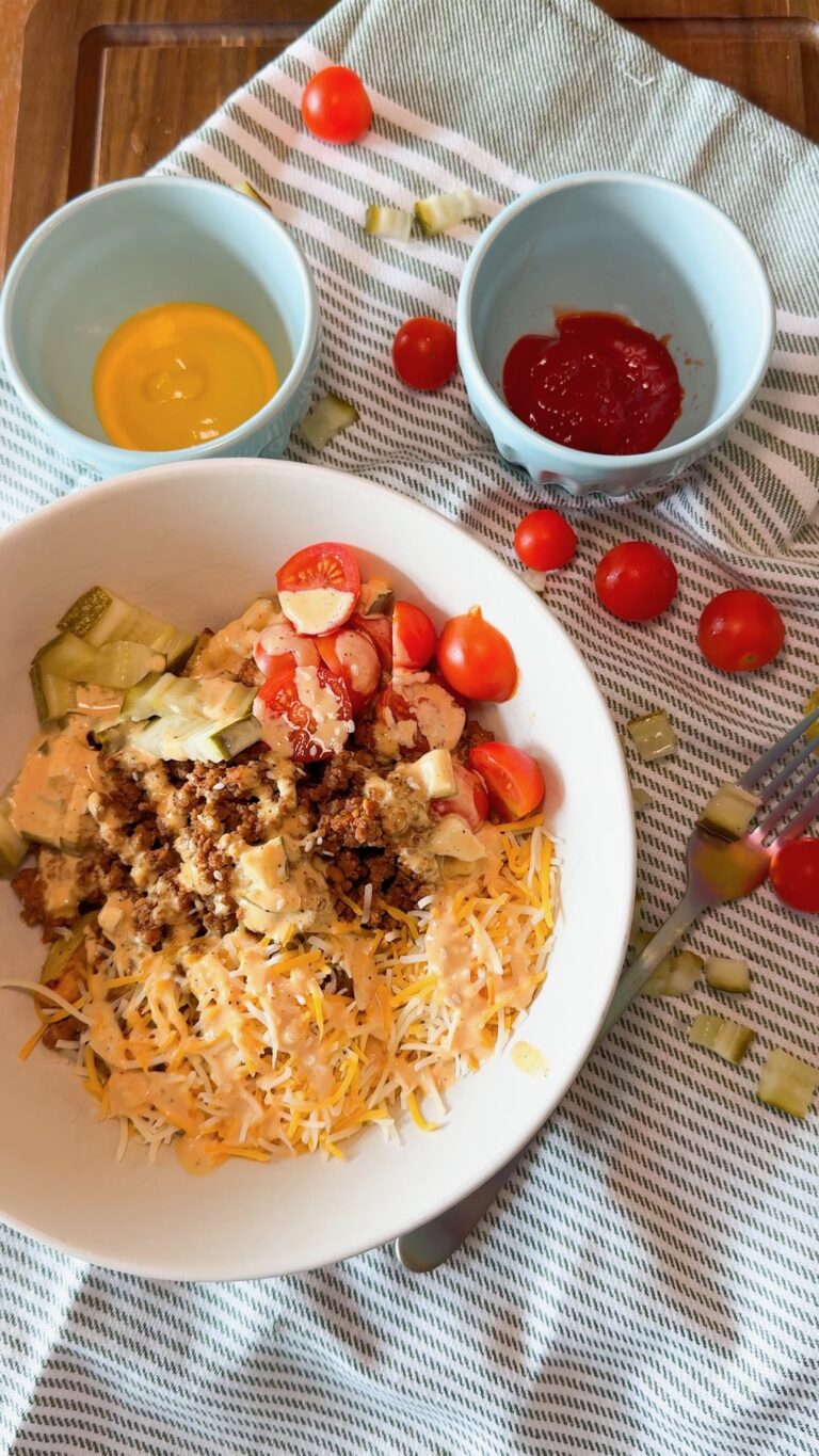 A white bowl with shredded cheese, ground beef, diced pickles, chopped lettuce, cherry tomatoes, and sauce sits on a striped cloth near bowls of ketchup and mustard, with scattered cherry tomatoes and pickles.