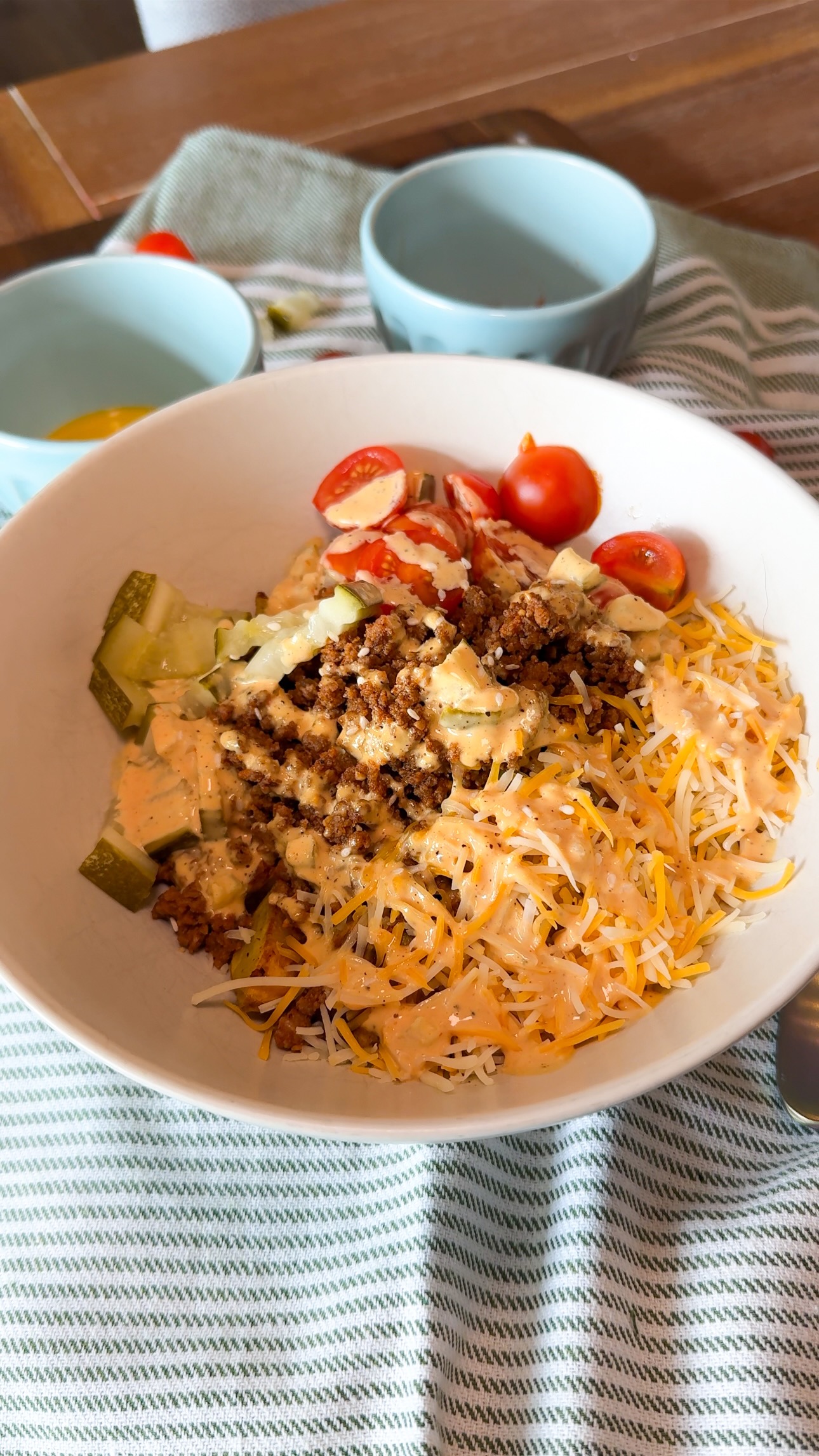 A bowl with shredded cheese, cherry tomatoes, pickles, ground meat, and sauce sits on a striped cloth, with a fork and two small bowls nearby.