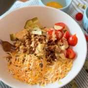 A bowl with shredded cheese, cherry tomatoes, pickles, ground meat, and sauce sits on a striped cloth, with a fork and two small bowls nearby.