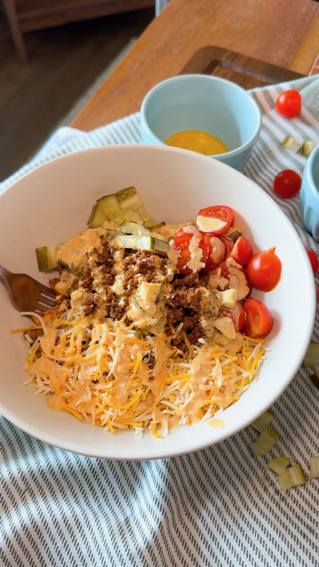A bowl with shredded cheese, cherry tomatoes, pickles, ground meat, and sauce sits on a striped cloth, with a fork and two small bowls nearby.