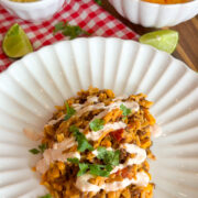A plate of Mexican-inspired rice with corn, ground meat, and sauce, garnished with cilantro, sits on a white plate. Nearby are bowls of shredded cheese, dry orzo, a sliced avocado, and lime wedges on a red plaid cloth.