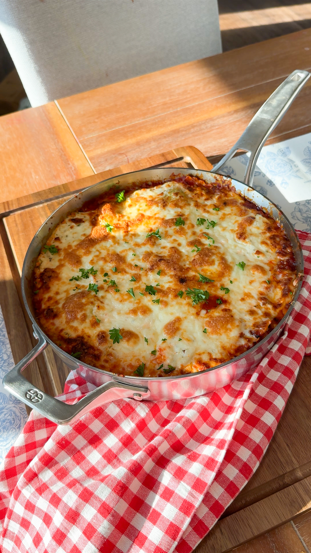 A serving of lasagna topped with parsley sits on a white plate, placed on a red checkered cloth. An onion, a small bowl, and parsley are on a wooden board in the background.
