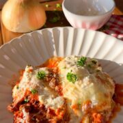 Two pieces of lasagna topped with melted cheese and herbs on a white plate. In the background, there’s a whole onion, a small bowl, chopped herbs, and a red-checkered cloth on a wooden surface.