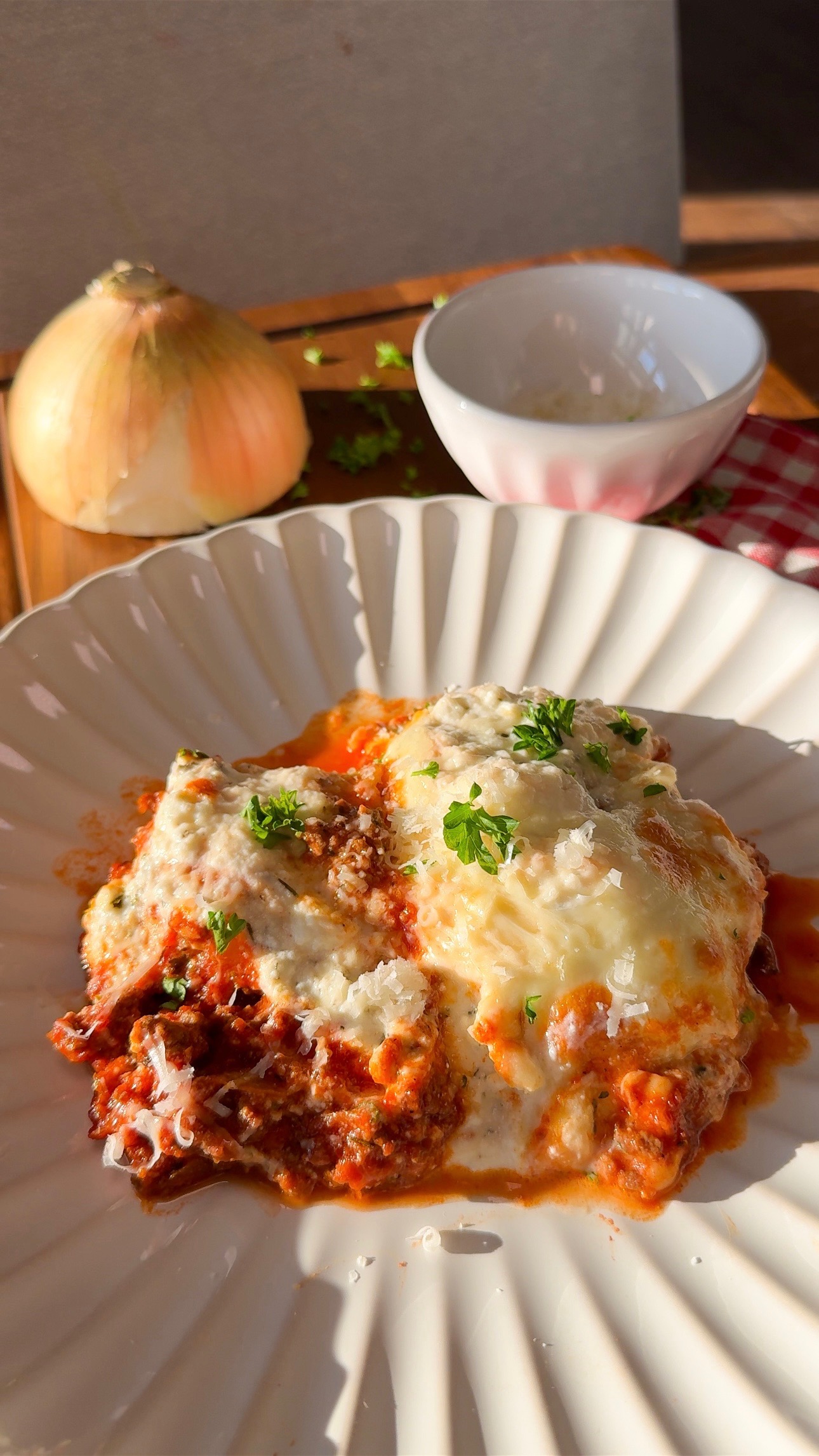 Two pieces of lasagna topped with melted cheese and herbs on a white plate. In the background, there’s a whole onion, a small bowl, chopped herbs, and a red-checkered cloth on a wooden surface.