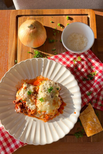 A plate of lasagna topped with herbs sits on a white scalloped plate. Nearby are a whole onion, a bowl of grated cheese, a block of cheese, and a red-checkered cloth on a wooden tray.