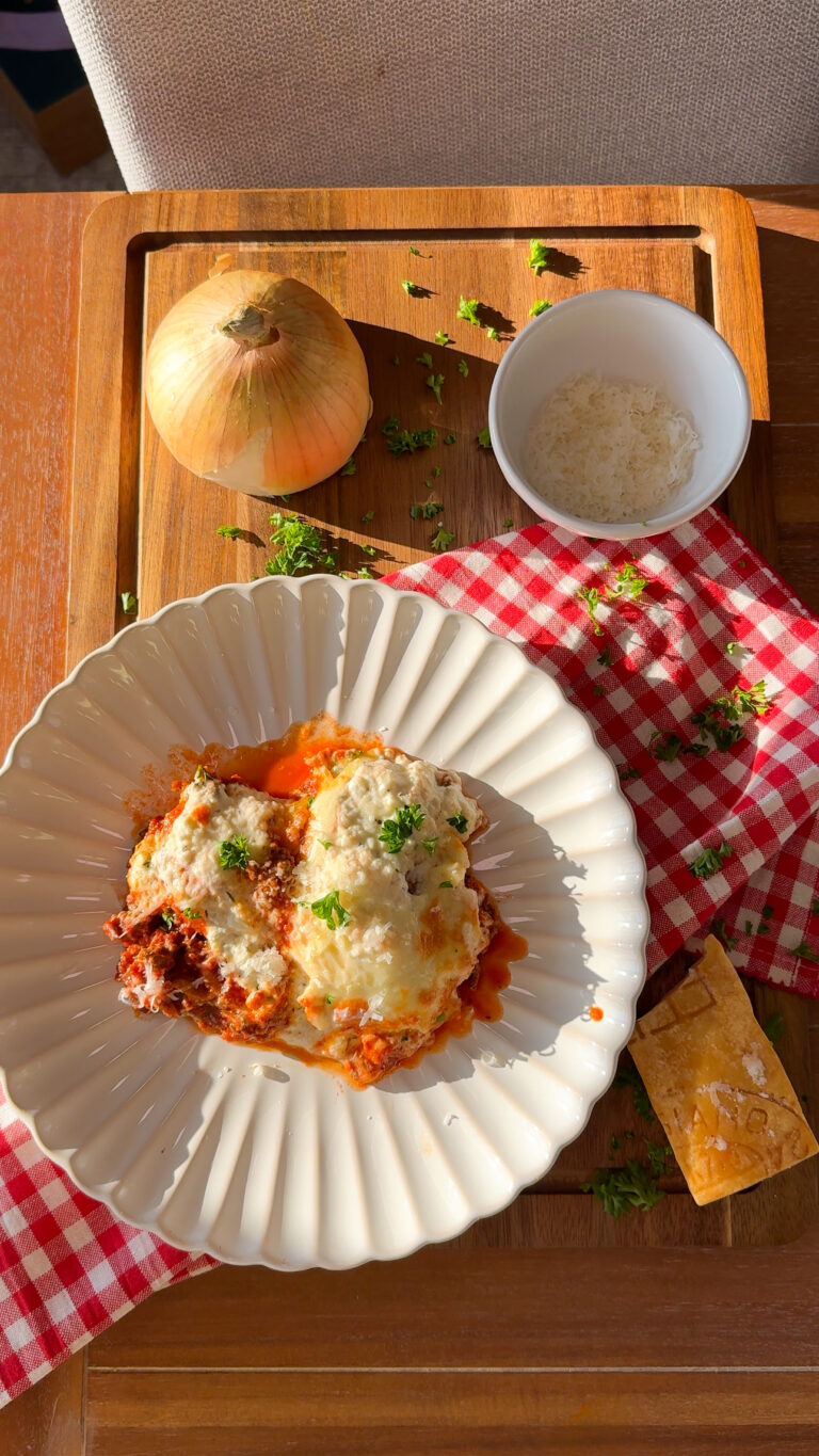 A plate of lasagna topped with herbs sits on a white scalloped plate. Nearby are a whole onion, a bowl of grated cheese, a block of cheese, and a red-checkered cloth on a wooden tray.