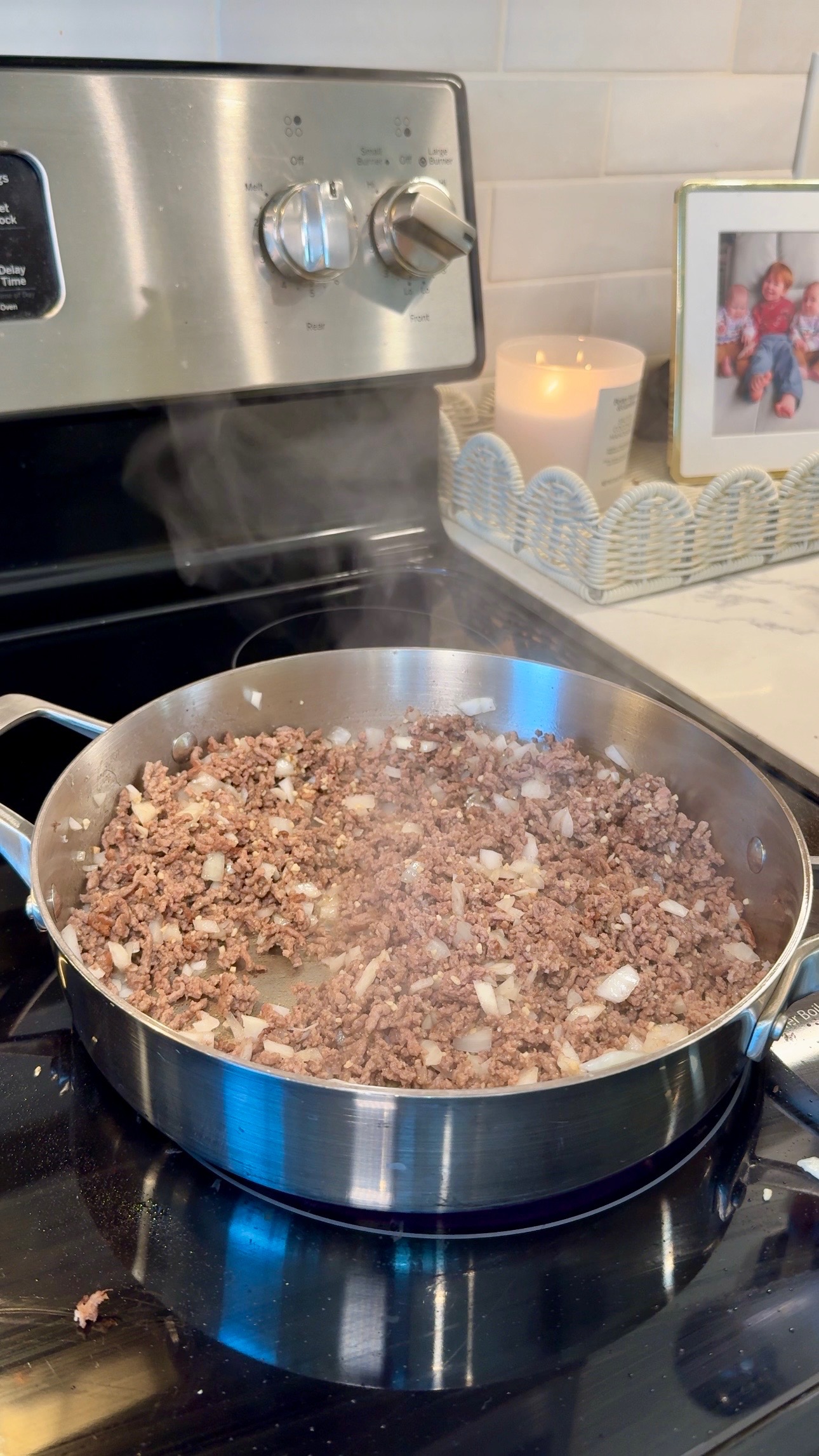 Ground beef and chopped onions cooking in a stainless steel pan on a stove. Steam rises from the pan. A lit candle and a framed photo sit on the counter in the background.