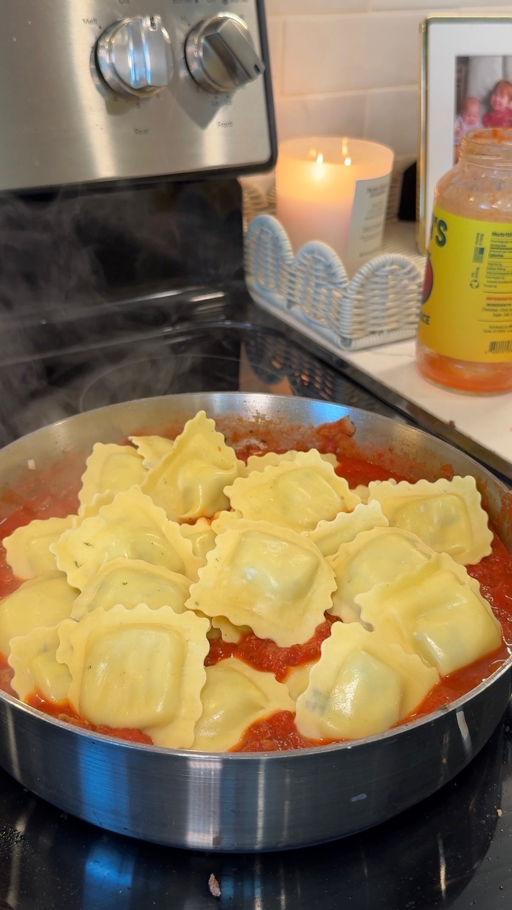 A pan of square ravioli cooks in red tomato sauce on a stovetop. Steam rises, and a candle, photo frame, and a jar of sauce are visible in the background.