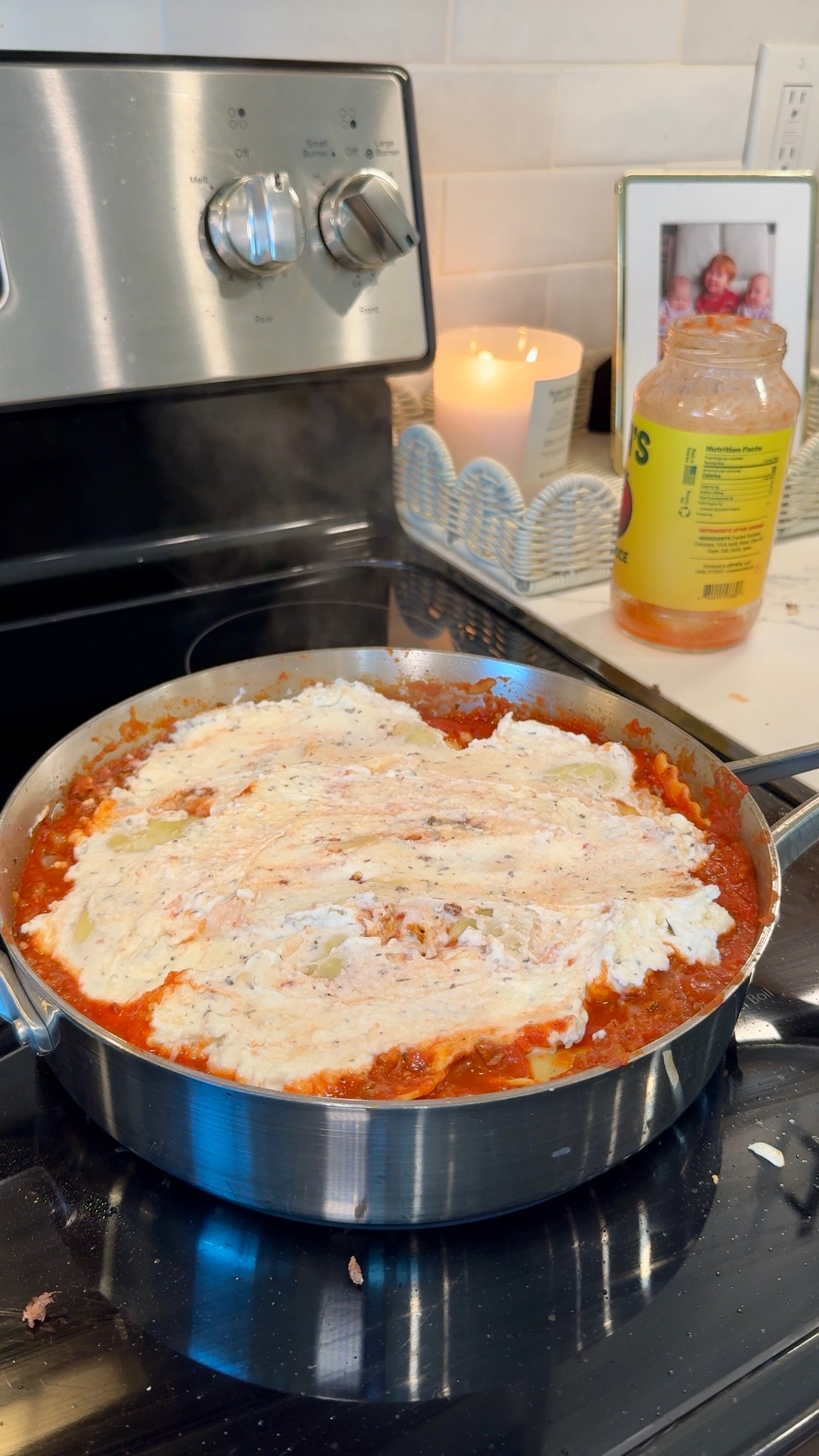 A large pan of lasagna is cooking on a stovetop. The cheese is melting on top of the tomato sauce. A candle, an open jar of pasta sauce, and a framed photo are on the counter in the background.