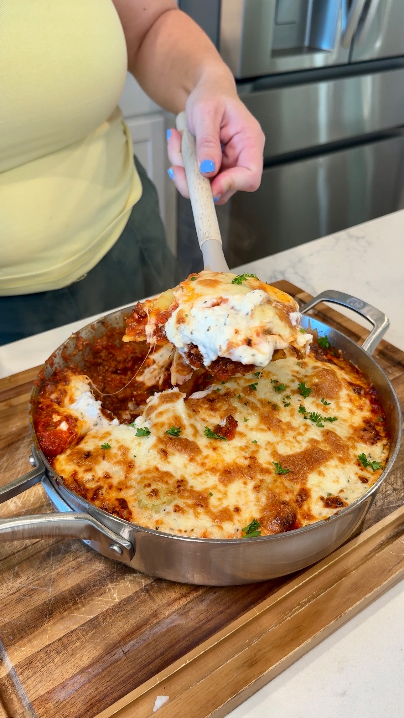 A person serves a cheesy, baked pasta dish from a skillet. The dish is topped with melted cheese and parsley, and sits on a wooden board in a kitchen setting.