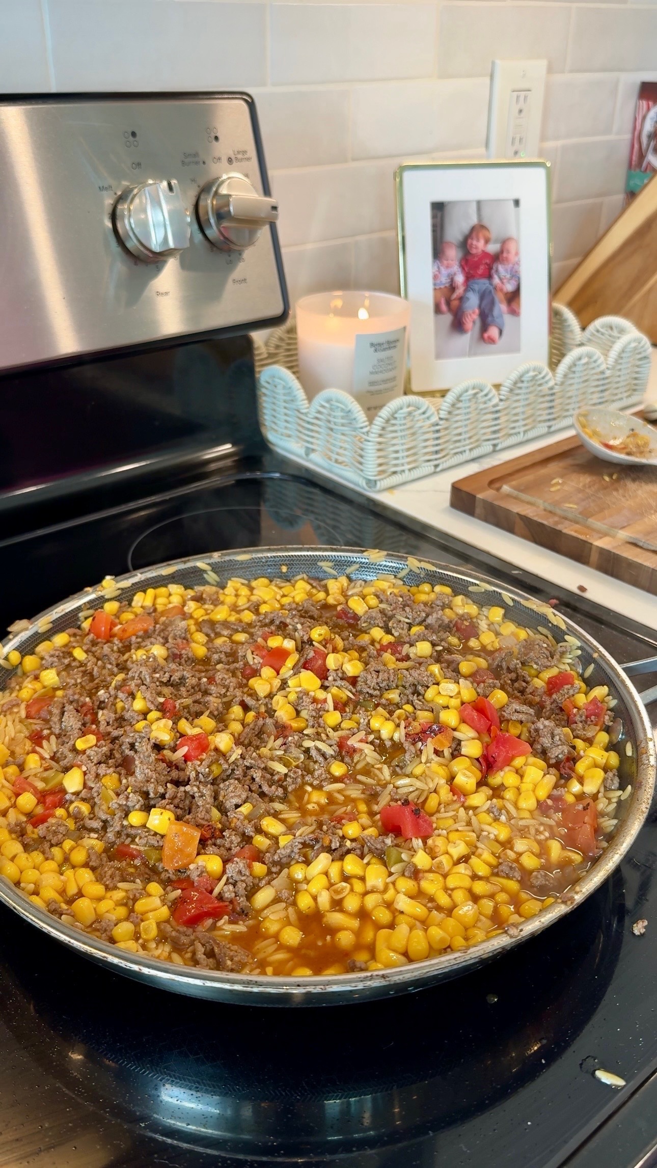A skillet on a stovetop contains a mixture of ground meat, corn, tomatoes, and peppers. In the background are a candle, a photo frame, and a cutting board on a kitchen counter.