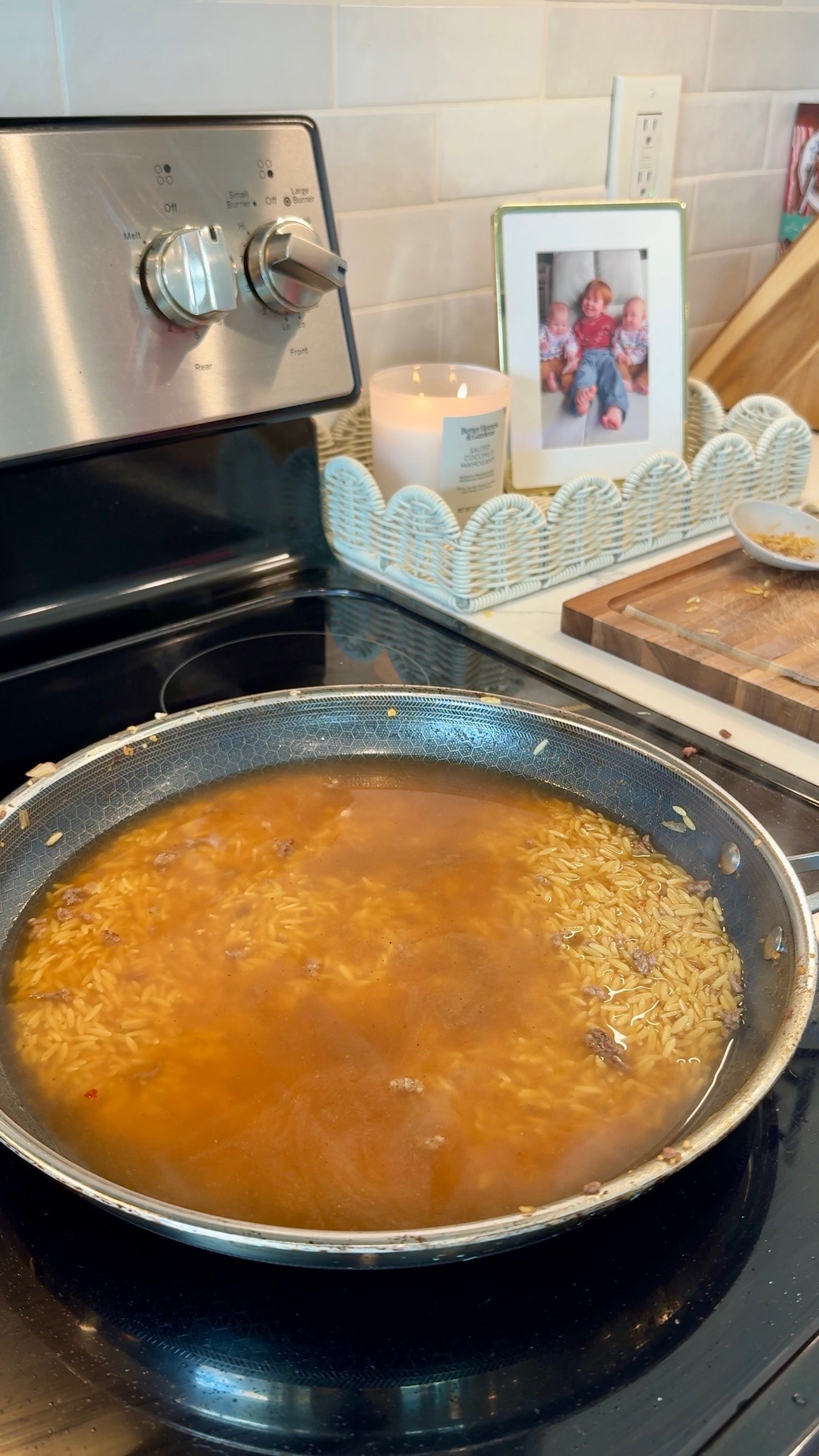 A pan of rice and broth simmers on a stove. Nearby are a lit candle, a framed family photo, and a cutting board with crumbs. The kitchen has a light, homey décor.