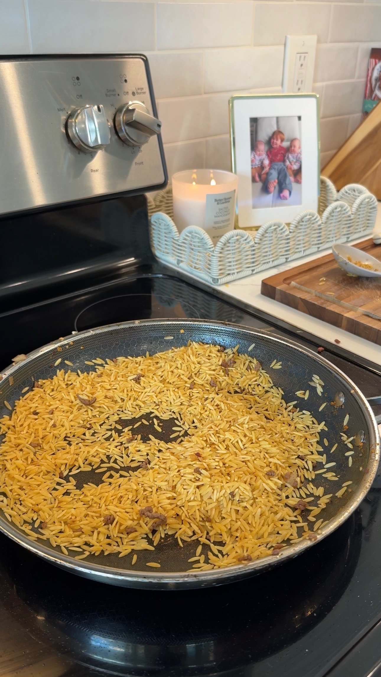 A pan of toasted orzo sits on a stovetop. Behind it are a candle, a framed family photo, and a small cutting board with kitchen items on a counter.