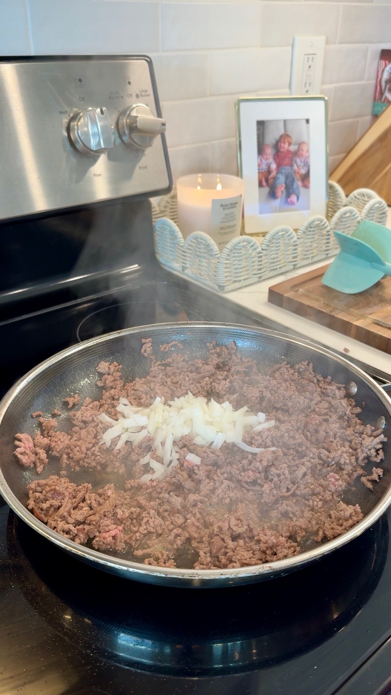 Ground beef and chopped onions cooking in a skillet on a stove. Steam rises from the pan. In the background are a lit candle, framed photo, and kitchen utensils on a countertop.