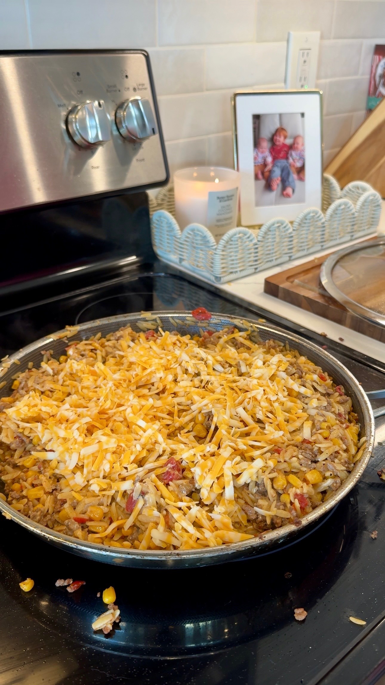 A skillet filled with a cheesy rice, beef, and vegetable mixture sits on a stovetop. In the background are a candle, a framed family photo, and kitchen decor.