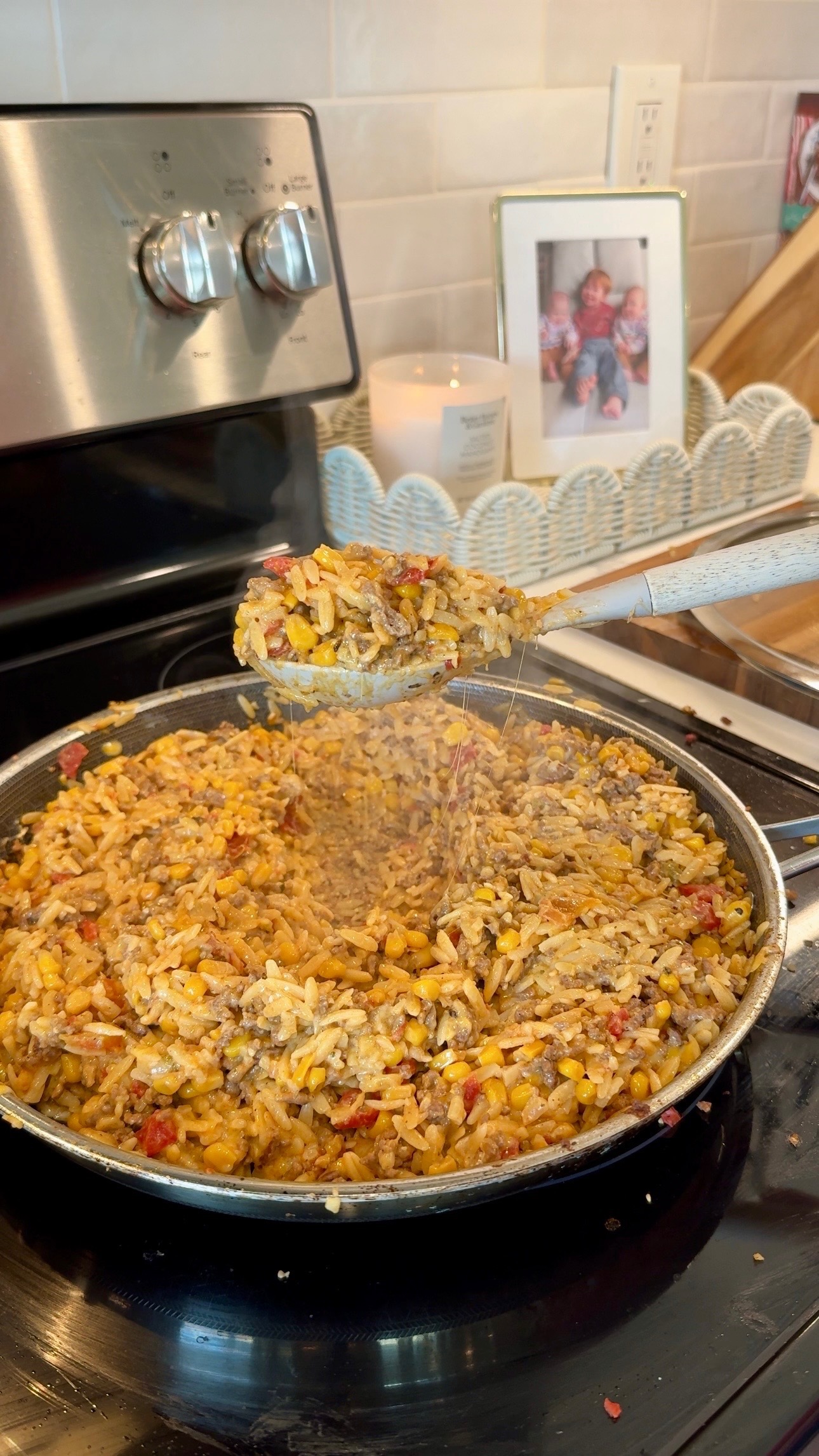 A skillet of cheesy rice, corn, and ground meat mixture cooking on a stove, with a spoon lifting some. A candle and framed photo sit on the counter in the background.