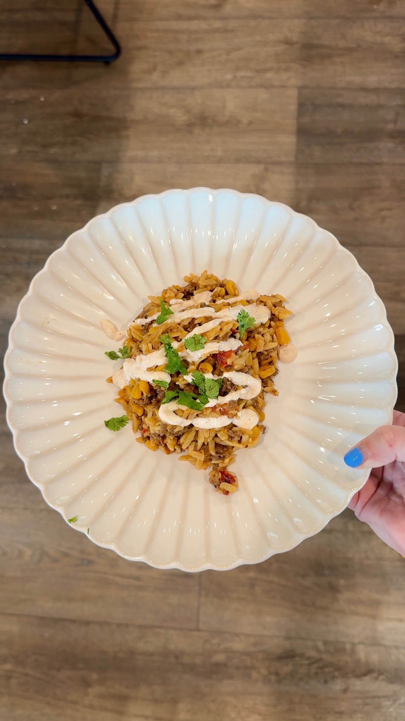 A hand with blue nail polish holds a scalloped white plate with a serving of seasoned rice, beans, veggies, and herbs, topped with a creamy white sauce and garnished with fresh cilantro.