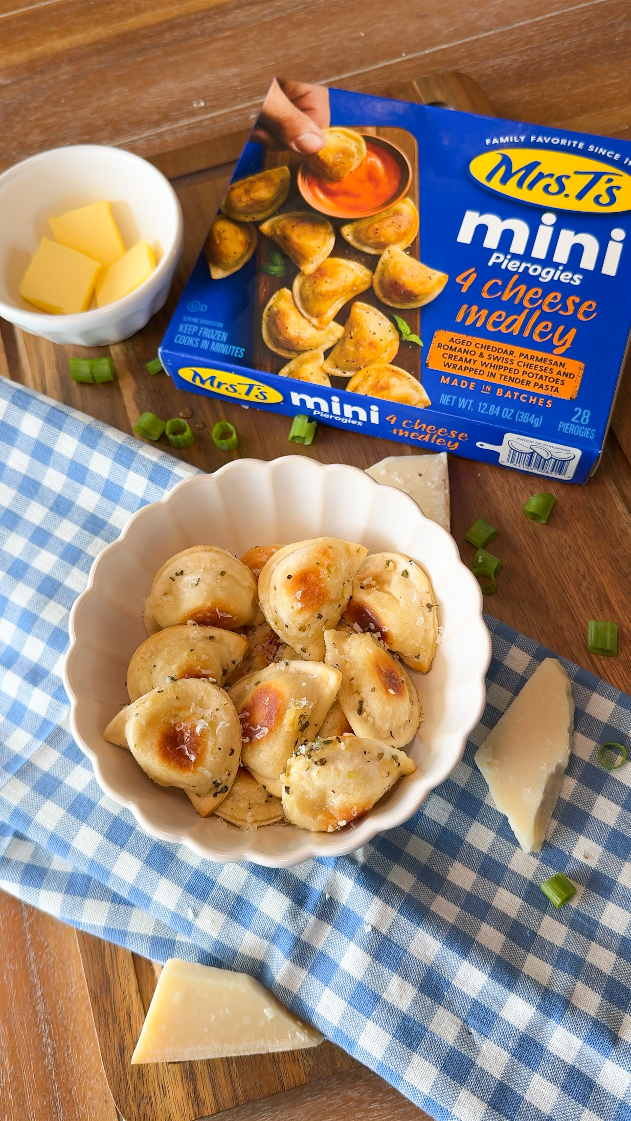 Bowl of cooked mini pierogies on a blue-checkered cloth, garnished with herbs. Nearby are sliced green onions, a dish of butter, cheese wedges, and a Mrs. T’s Mini Pierogies 4 Cheese Medley box.