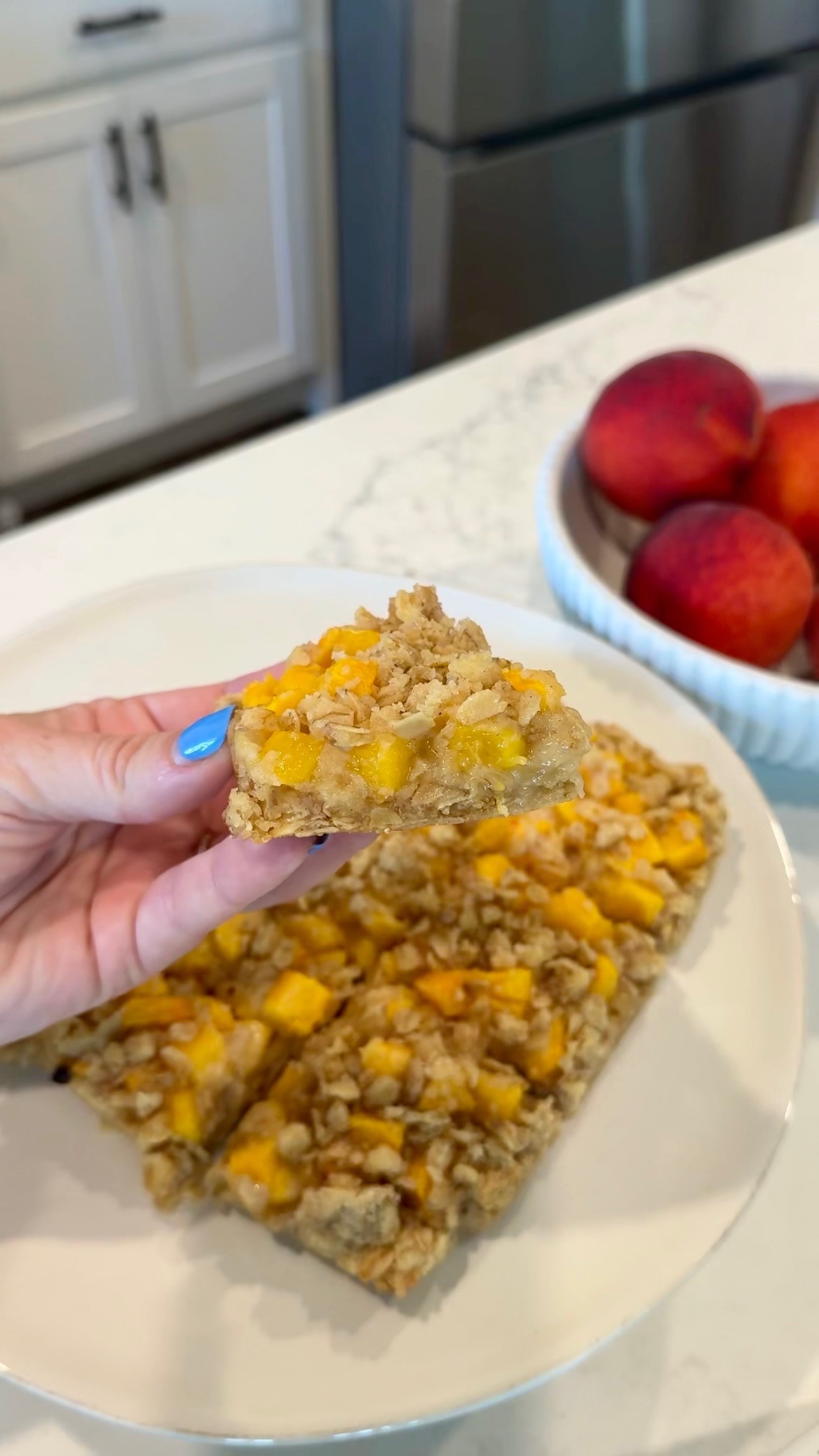 A hand holding a peach crumb bar with oat topping above a plate of more bars; a bowl of fresh peaches sits in the background on a marble countertop.