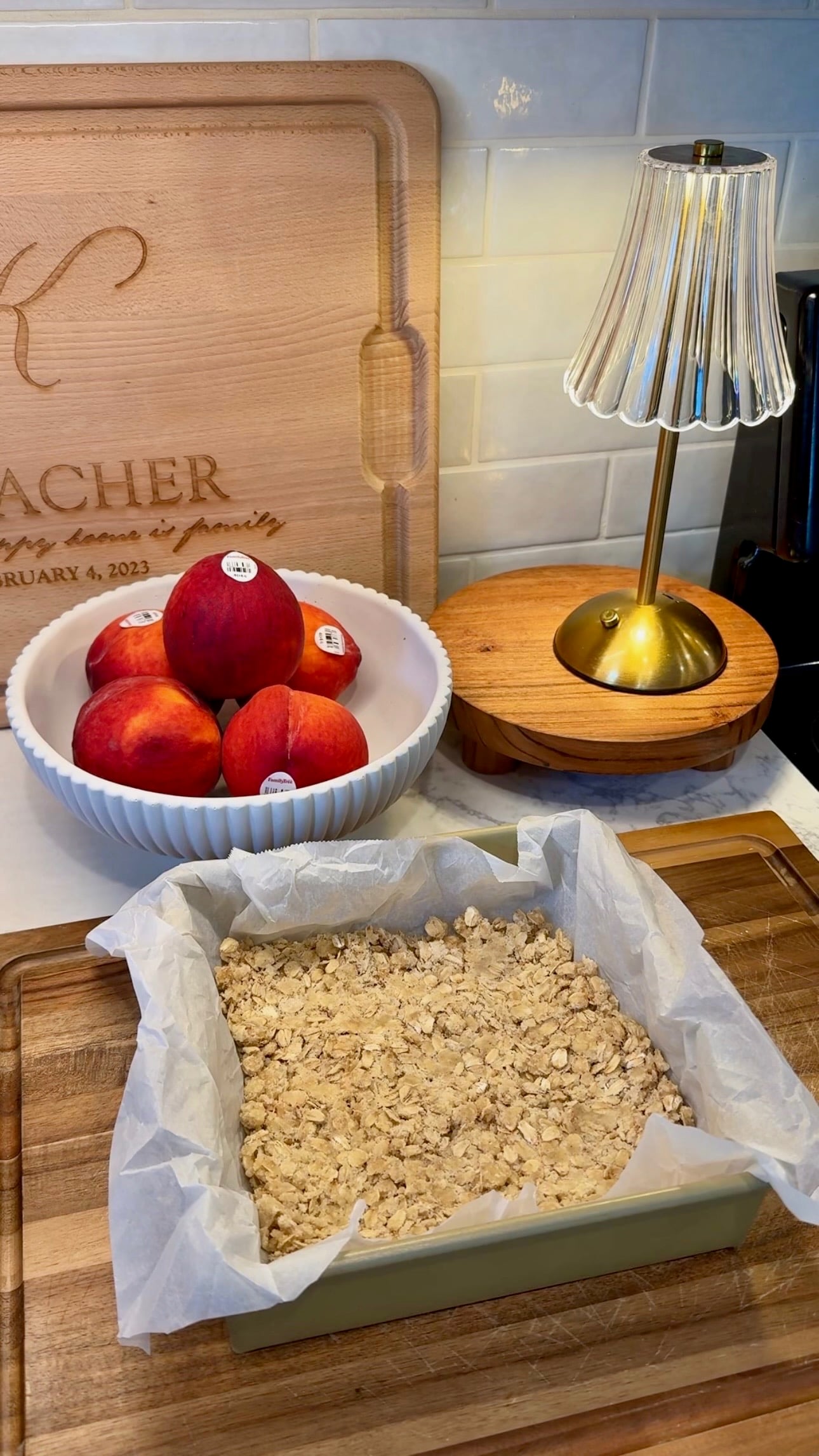 A parchment-lined pan with oat crumble sits on a wooden board. Behind it is a bowl of apples, a small lamp, and a wooden cutting board engraved with text. The scene is set on a kitchen counter.