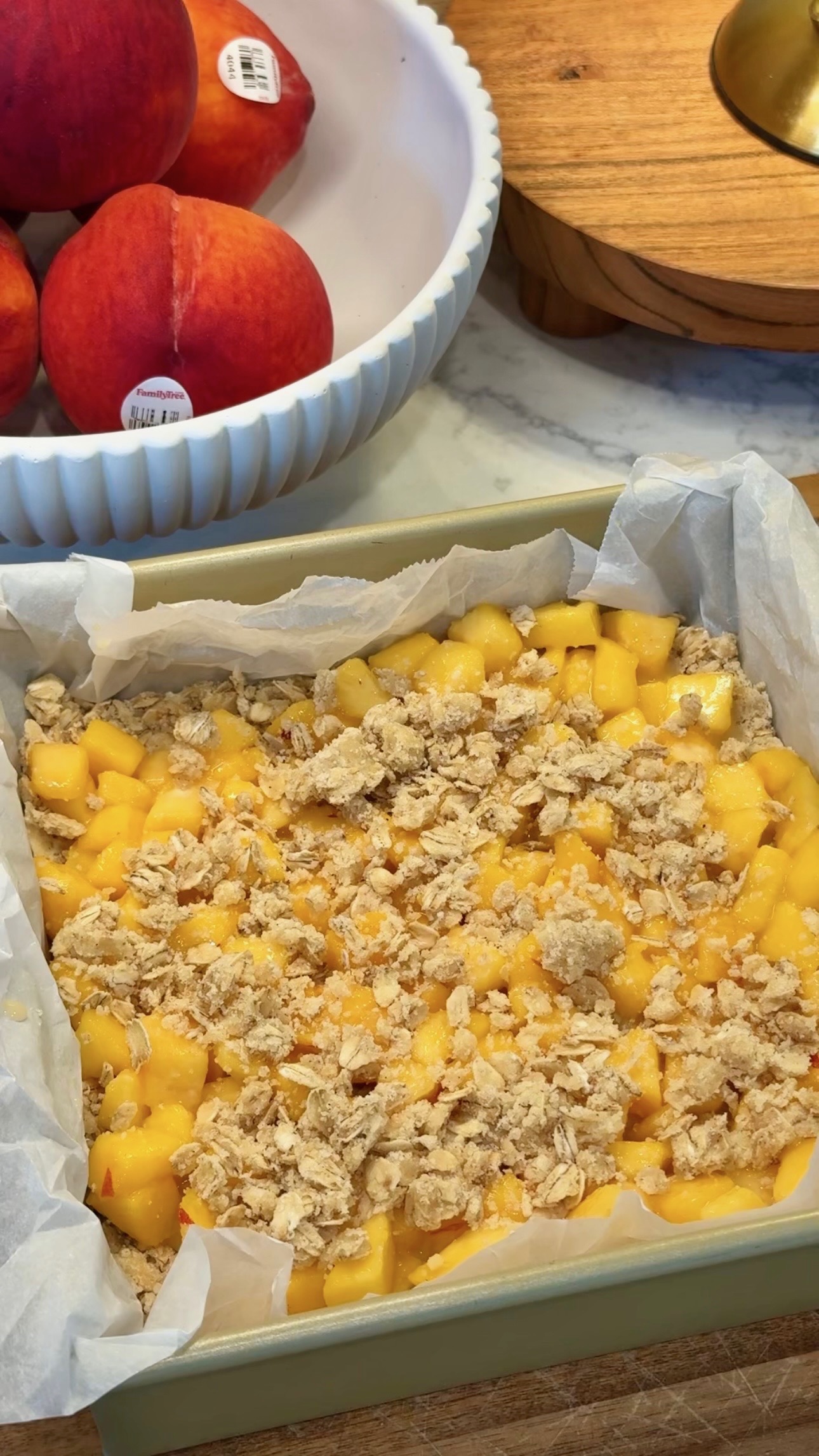 A baking pan lined with parchment paper, filled with diced peaches and topped with a crumbly oat mixture, sits on a marble counter. A bowl of whole peaches is in the background.