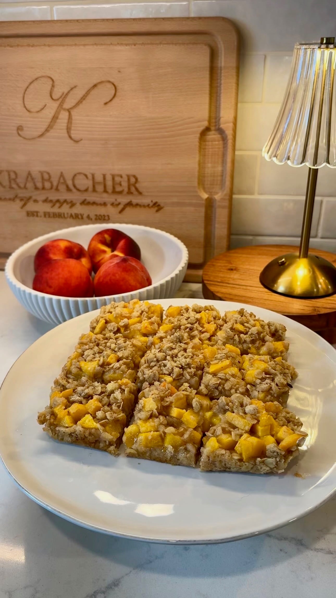 A plate of peach crumble bars sits on a counter near a bowl of fresh peaches, a small lamp, and a personalized wooden cutting board with engraved text in the background.