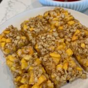 A plate with three oat and fruit bars, featuring visible chunks of yellow fruit, sits on a marble counter. In the background, a bowl filled with red and orange fruit is partially visible.