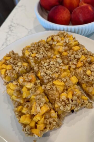 A plate with three oat and fruit bars, featuring visible chunks of yellow fruit, sits on a marble counter. In the background, a bowl filled with red and orange fruit is partially visible.