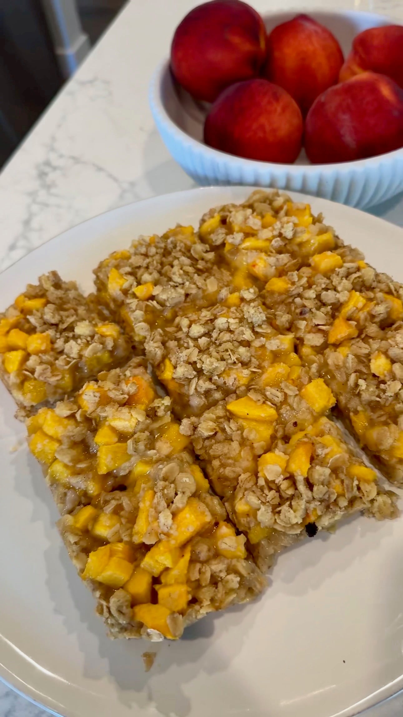 A plate with three oat and fruit bars, featuring visible chunks of yellow fruit, sits on a marble counter. In the background, a bowl filled with red and orange fruit is partially visible.