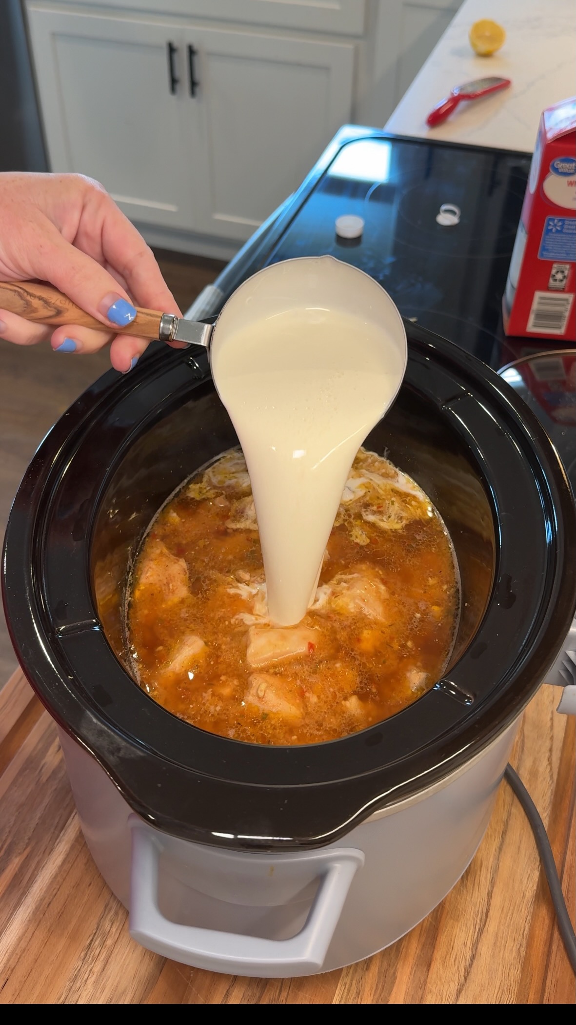 A hand pours a cup of cream into a slow cooker filled with seasoned chicken and broth, set on a kitchen countertop.